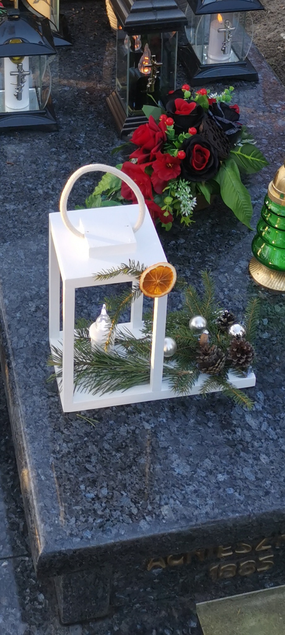 Dekoracja nagrobna: biały lampion z suszoną pomarańczą, gałązkami świerku, szyszkami i srebrnymi bombkami, na ciemnym, granitowym nagrobku z kwiatami i lampionami.