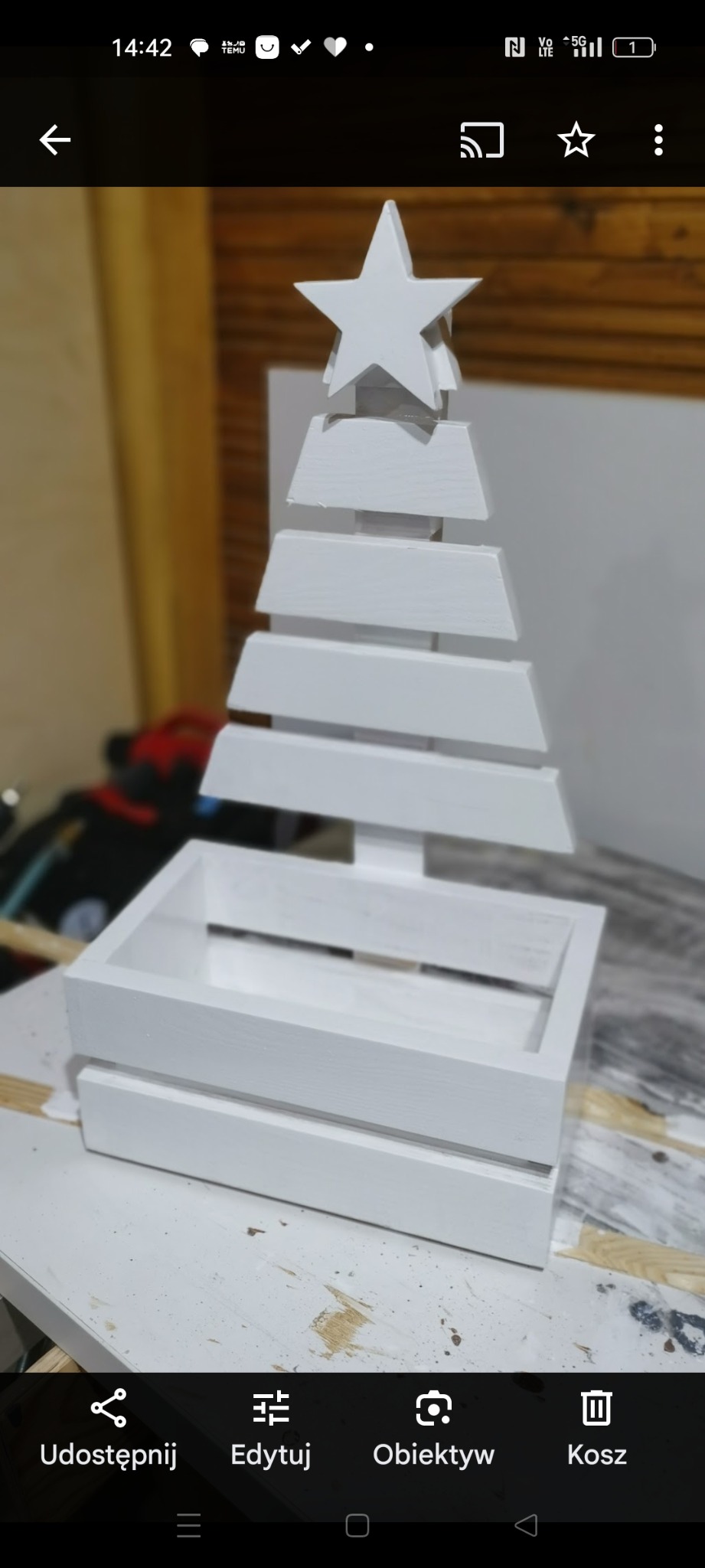 Biała, drewniana choinka dekoracyjna z gwiazdą na szczycie, umieszczona w skrzynce. Widoczne ślady farby, sugerujące ręczne wykonanie.