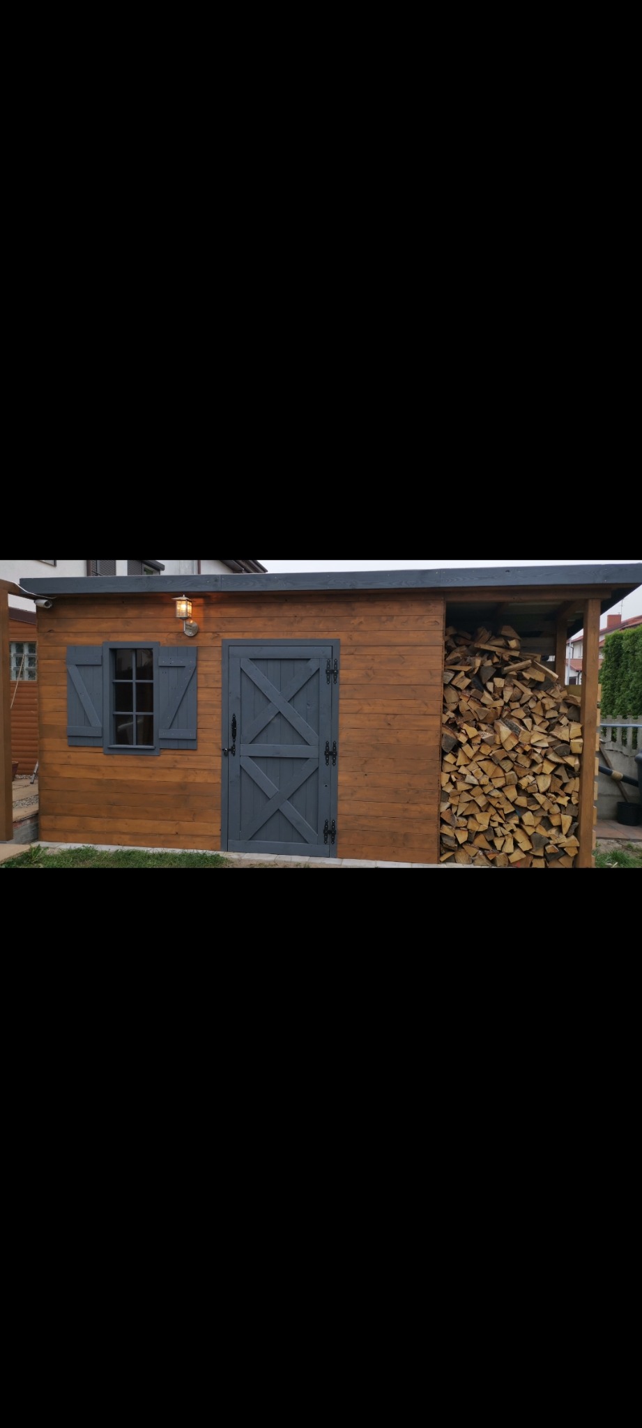 Drewniana drewutnia z szarą stolarką, oświetlona lampą. Z boku stos drewna. Widoczne szare drzwi z krzyżowym wzorem i okienko z okiennicą.