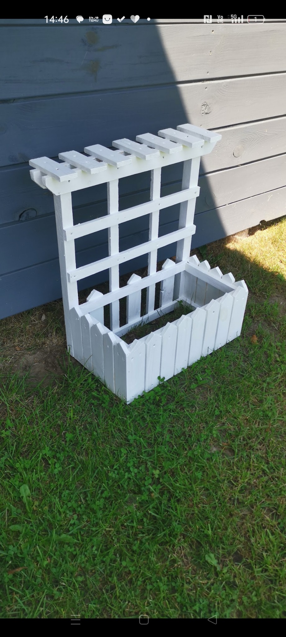 Biała, drewniana konstrukcja ogrodowa z kratką i donicą, ustawiona na trawie przy szarej ścianie. Widoczne detale wykonania i świeża zieleń trawy.