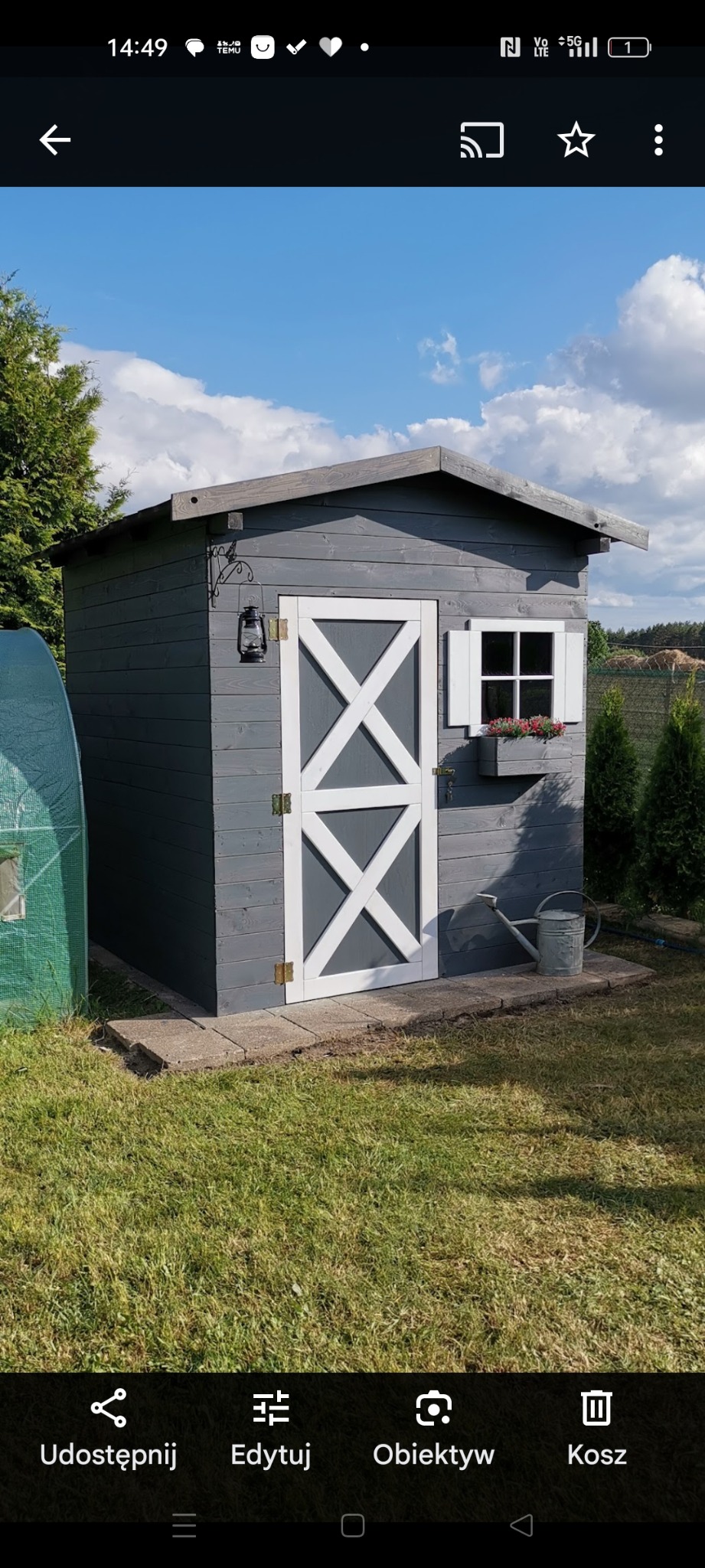 Szary, drewniany domek narzędziowy z białymi drzwiami i okiennicą, udekorowany lampionem i kwiatami, stoi na trawniku obok szklarni i iglaków.