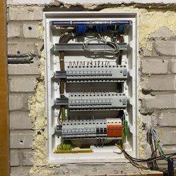 GRZEGORZ BYKOWSKI GANC-INSTALATOR - Rozdzielnica elektryczna w trakcie montażu w ścianie z cegły, zabezpieczona pianką montażową. Widoczne przewody i elementy instalacji.