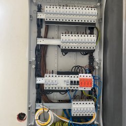 GRZEGORZ BYKOWSKI GANC-INSTALATOR - Rozdzielnica elektryczna w obudowie, widoczne bezpieczniki, przewody w różnych kolorach, listwy zaciskowe. Instalacja w trakcie montażu, widoczny otwór w ścianie.
