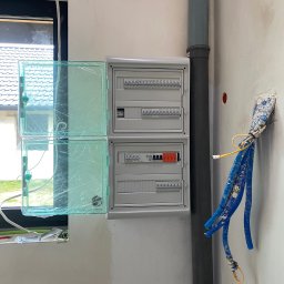 GRZEGORZ BYKOWSKI GANC-INSTALATOR - Rozdzielnia elektryczna z otwartymi, zabezpieczonymi folią drzwiczkami, widoczne przewody w ścianie i rura kanalizacyjna. Ściana w trakcie wykończenia.
