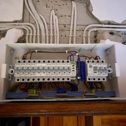 GRZEGORZ BYKOWSKI GANC-INSTALATOR - Otwarta skrzynka rozdzielcza z bezpiecznikami i przewodami, zamontowana na ścianie z widocznymi przewodami w bruzdach. Widoczne elementy instalacji elektrycznej.