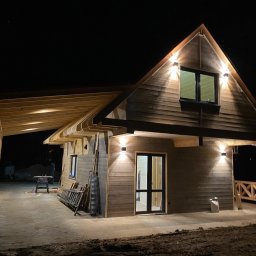 GRZEGORZ BYKOWSKI GANC-INSTALATOR - Nocny widok drewnianego domu z podświetleniem elewacji i zadaszonego tarasu. Widoczne lampy ścienne, okno i drzwi. Częściowo wykończony teren wokół budynku.