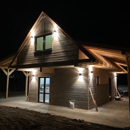 GRZEGORZ BYKOWSKI GANC-INSTALATOR - Nocny widok drewnianego domu z oświetleniem zewnętrznym. Lampy ścienne podkreślają architekturę budynku. Widoczne elementy konstrukcyjne dachu i tarasu.