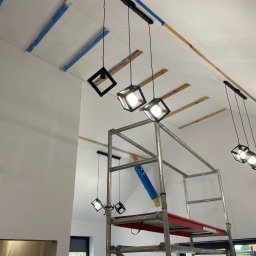 GRZEGORZ BYKOWSKI GANC-INSTALATOR - Montaż nowoczesnych lamp wiszących w surowym wnętrzu. Widoczne rusztowanie, zabezpieczenia malarskie i drewniane elementy konstrukcyjne. Oświetlenie w stylu industrialnym.