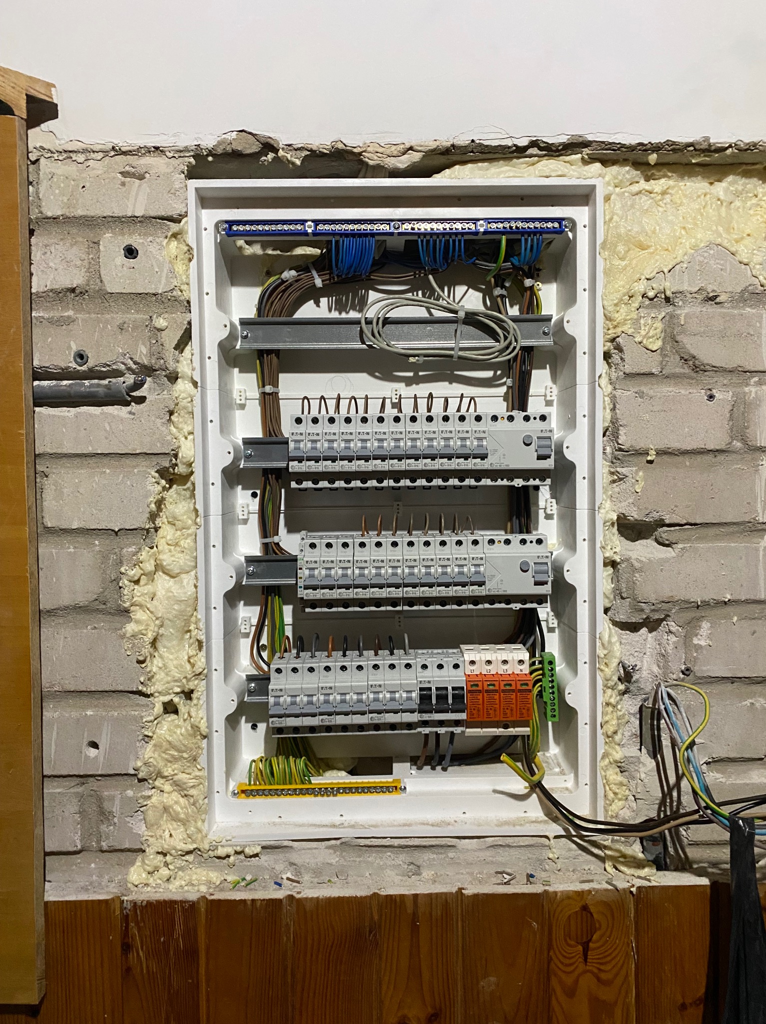 Rozdzielnica elektryczna w trakcie montażu w ścianie z cegły, zabezpieczona pianką montażową. Widoczne przewody i elementy instalacji.