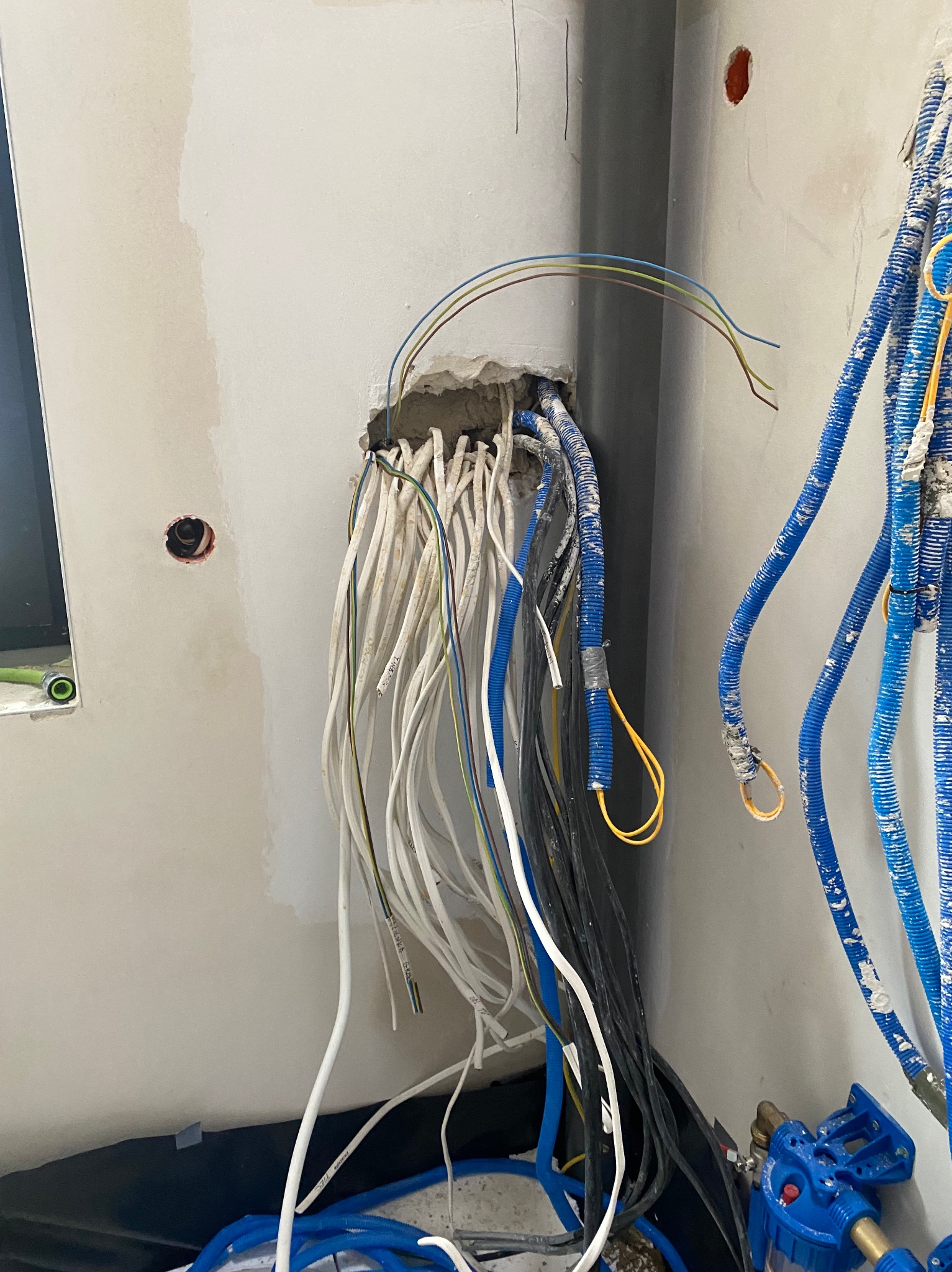 Wiązka przewodów elektrycznych wychodząca ze ściany, obok rur instalacyjnych w nieukończonym wnętrzu. Widoczne kable w różnych kolorach i grubościach.