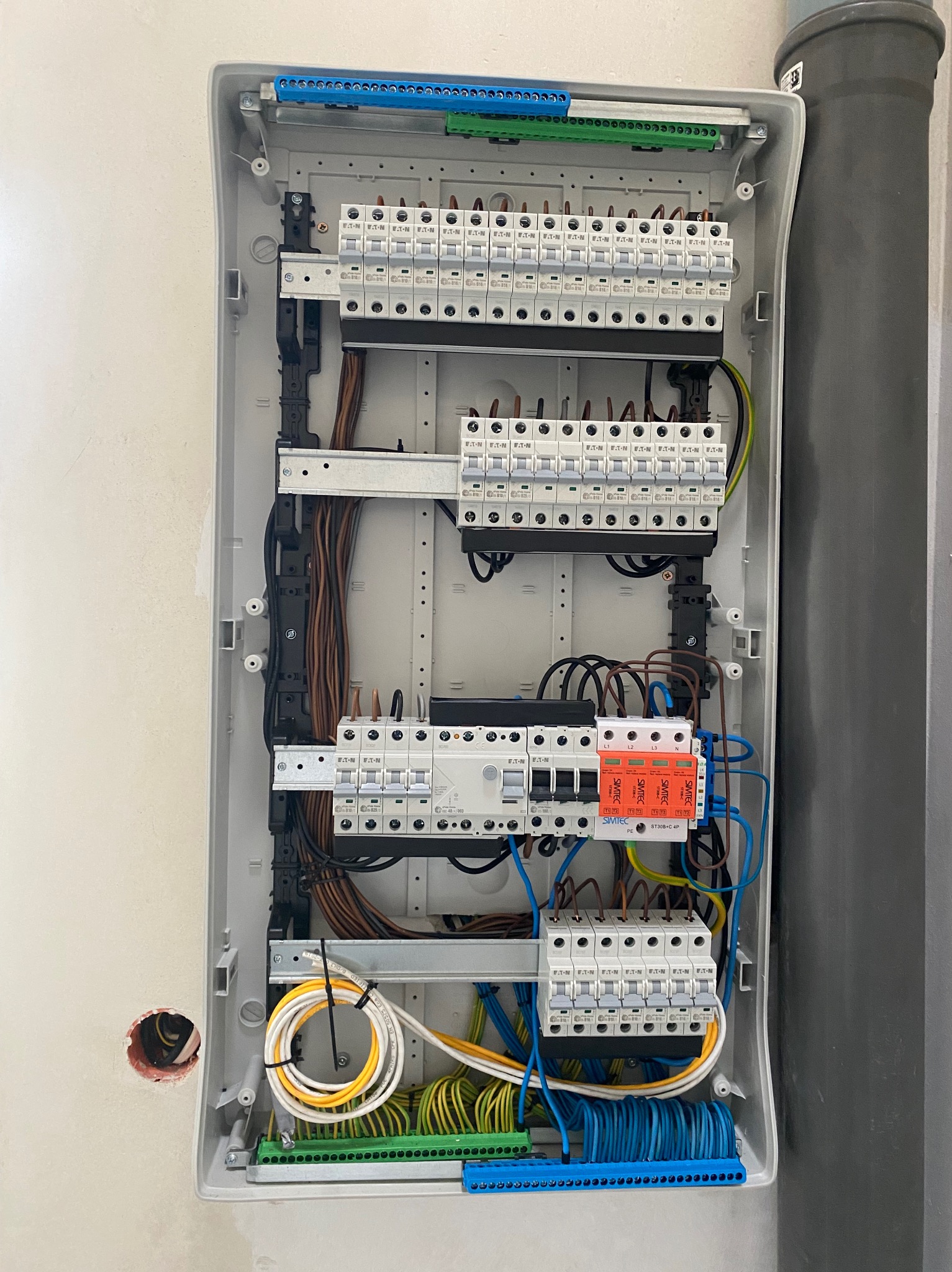 Rozdzielnica elektryczna w obudowie, widoczne bezpieczniki, przewody w różnych kolorach, listwy zaciskowe. Instalacja w trakcie montażu, widoczny otwór w ścianie.