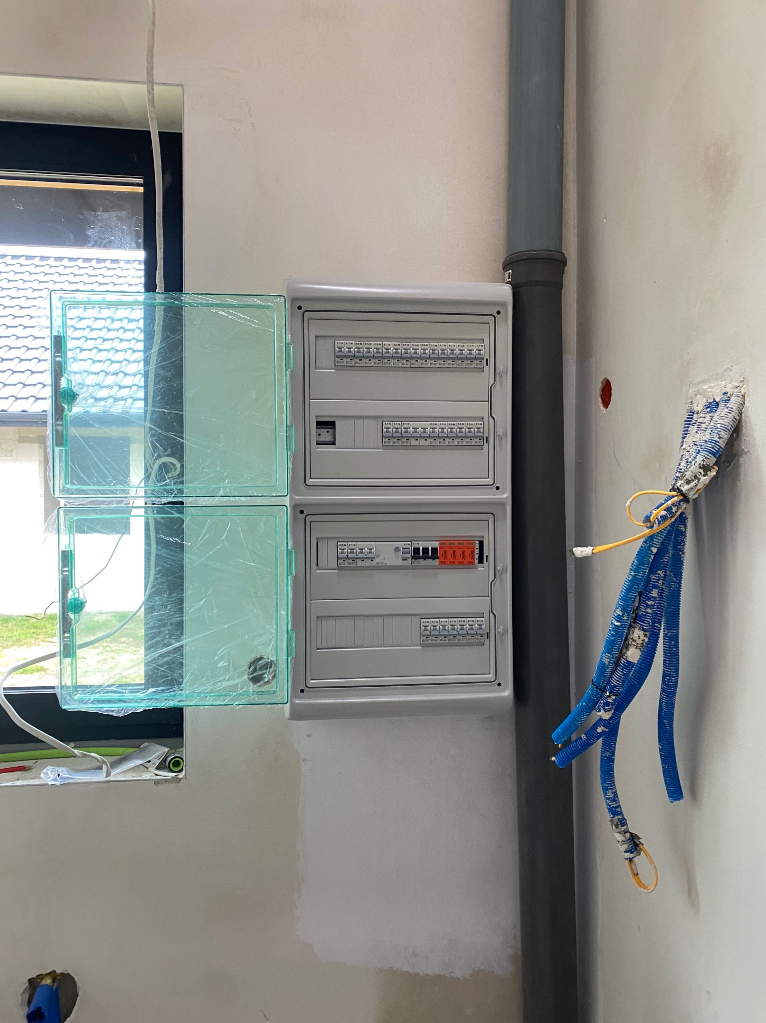 Rozdzielnia elektryczna z otwartymi, zabezpieczonymi folią drzwiczkami, widoczne przewody w ścianie i rura kanalizacyjna. Ściana w trakcie wykończenia.