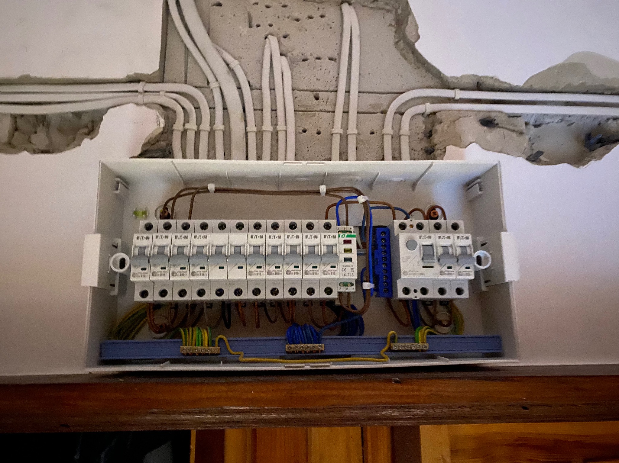 Otwarta skrzynka rozdzielcza z bezpiecznikami i przewodami, zamontowana na ścianie z widocznymi przewodami w bruzdach. Widoczne elementy instalacji elektrycznej.