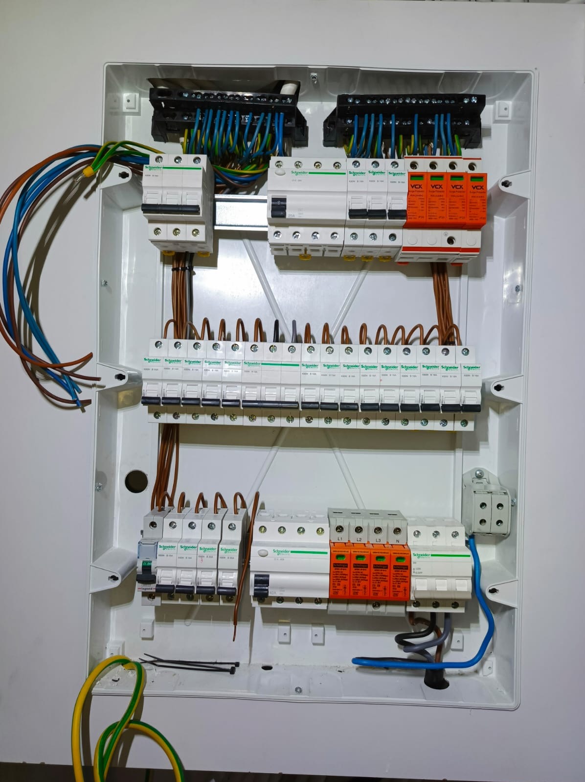 Rozdzielnica elektryczna w białej obudowie z widocznymi bezpiecznikami, przewodami i elementami zabezpieczeń. Widok z góry, oświetlenie naturalne.