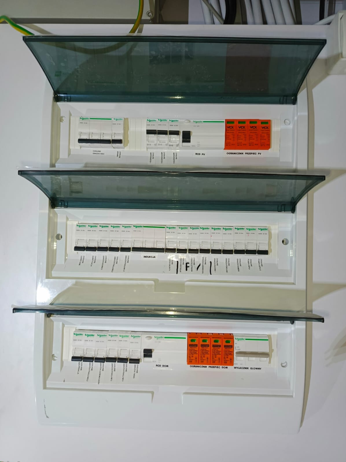 Rozdzielnica elektryczna z otwartymi klapami, widoczne zabezpieczenia różnicowoprądowe i ograniczniki przepięć. Instalacja wewnątrz budynku, białe tło.