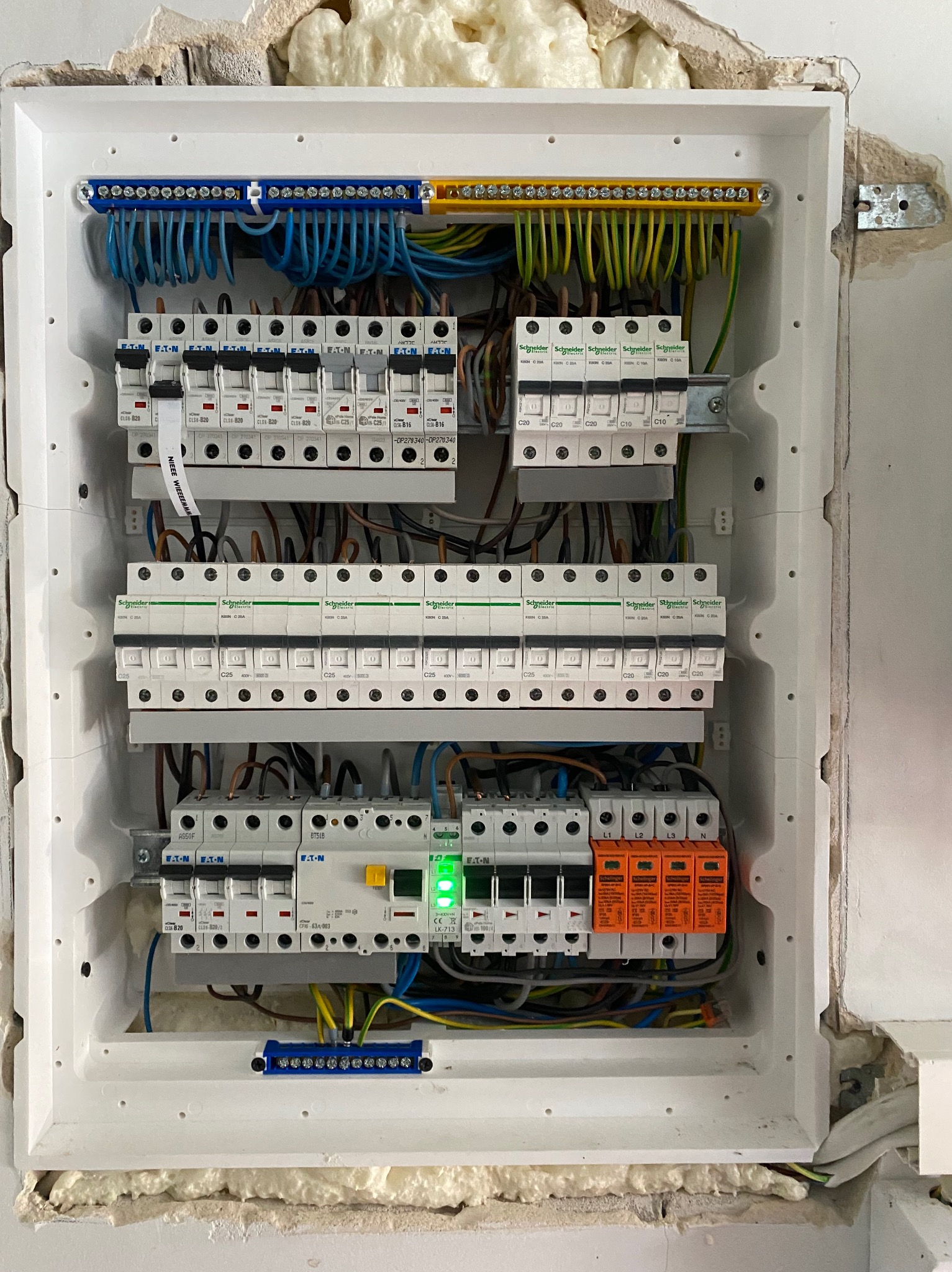 Rozdzielnica elektryczna z bezpiecznikami i przewodami w białej obudowie, zamontowana w ścianie z widoczną pianką montażową. Widoczne różne typy zabezpieczeń i oznaczenia.