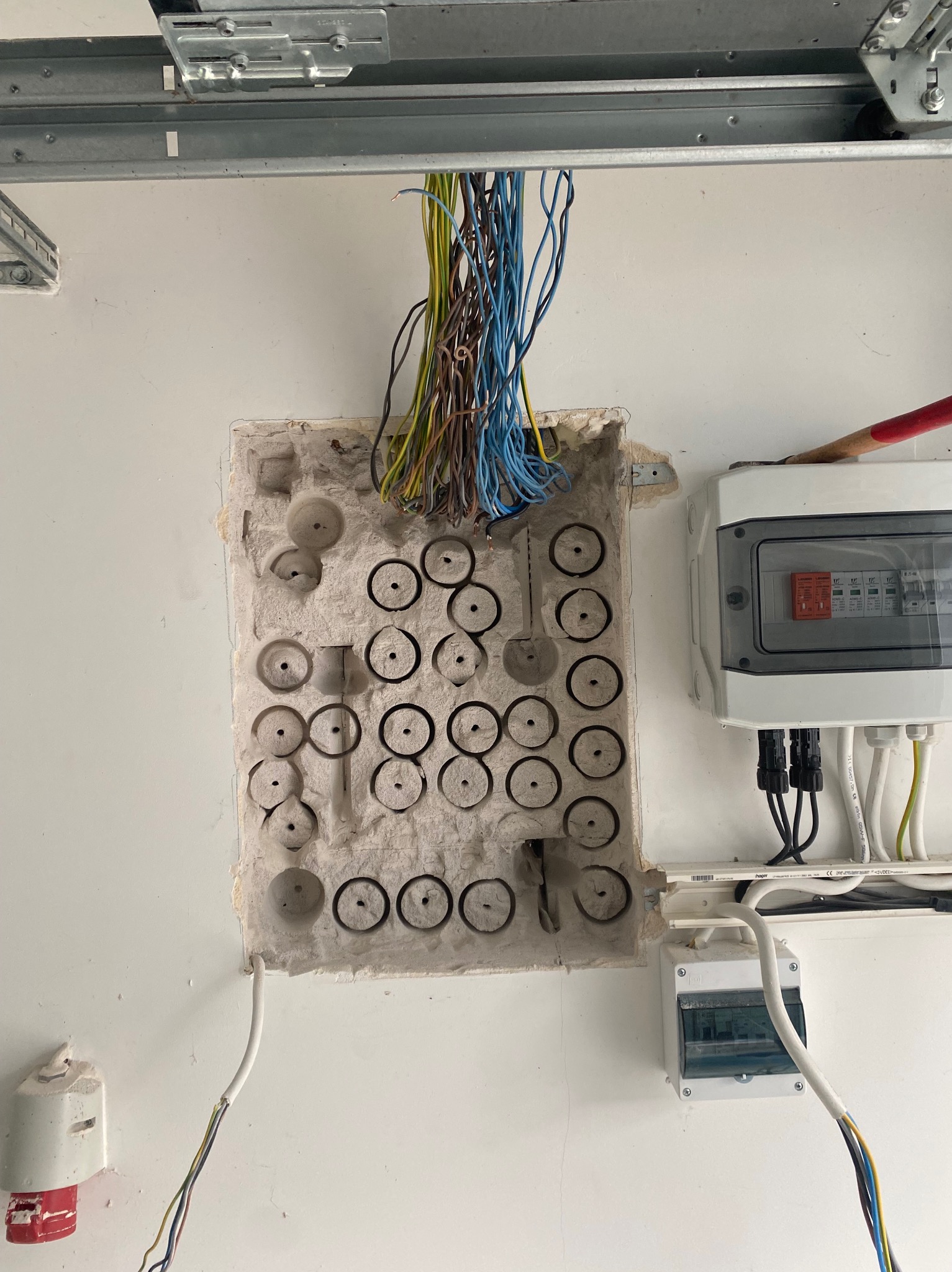 Otwór w ścianie z wystającymi przewodami, obok skrzynka elektryczna z bezpiecznikami. Widoczne kable w różnych kolorach. Etap prac.