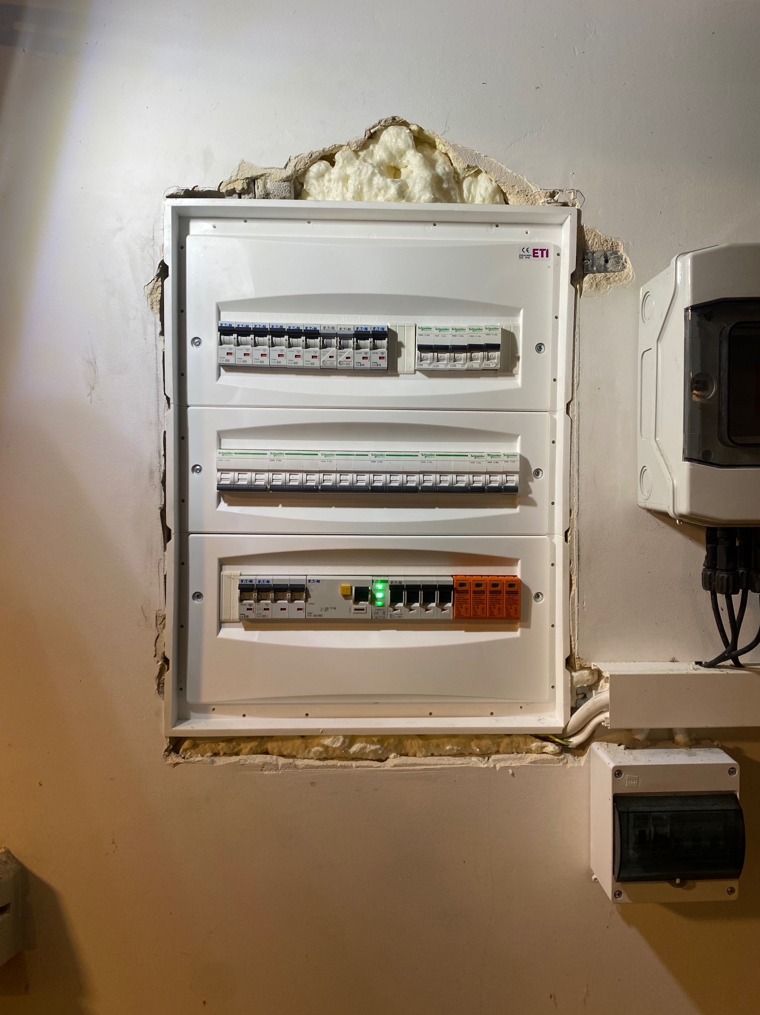 Rozdzielnica elektryczna ETI z zabezpieczeniami Eaton i Schneider, zamontowana w ścianie z widocznymi śladami montażu i pianką uszczelniającą. Widoczne przewody i dodatkowa skrzynka.