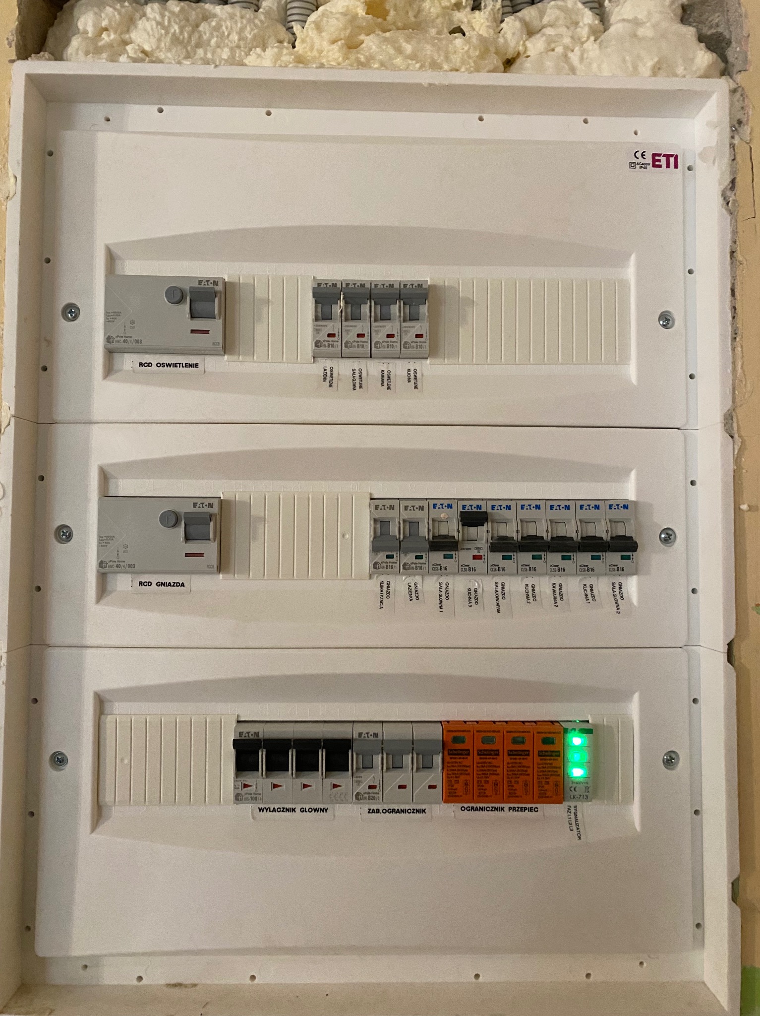 Rozdzielnica elektryczna z wyłącznikami Eaton i ogranicznikami przepięć, widoczne oznaczenia obwodów i sygnalizator faz, zamontowana w ścianie z pianką montażową.
