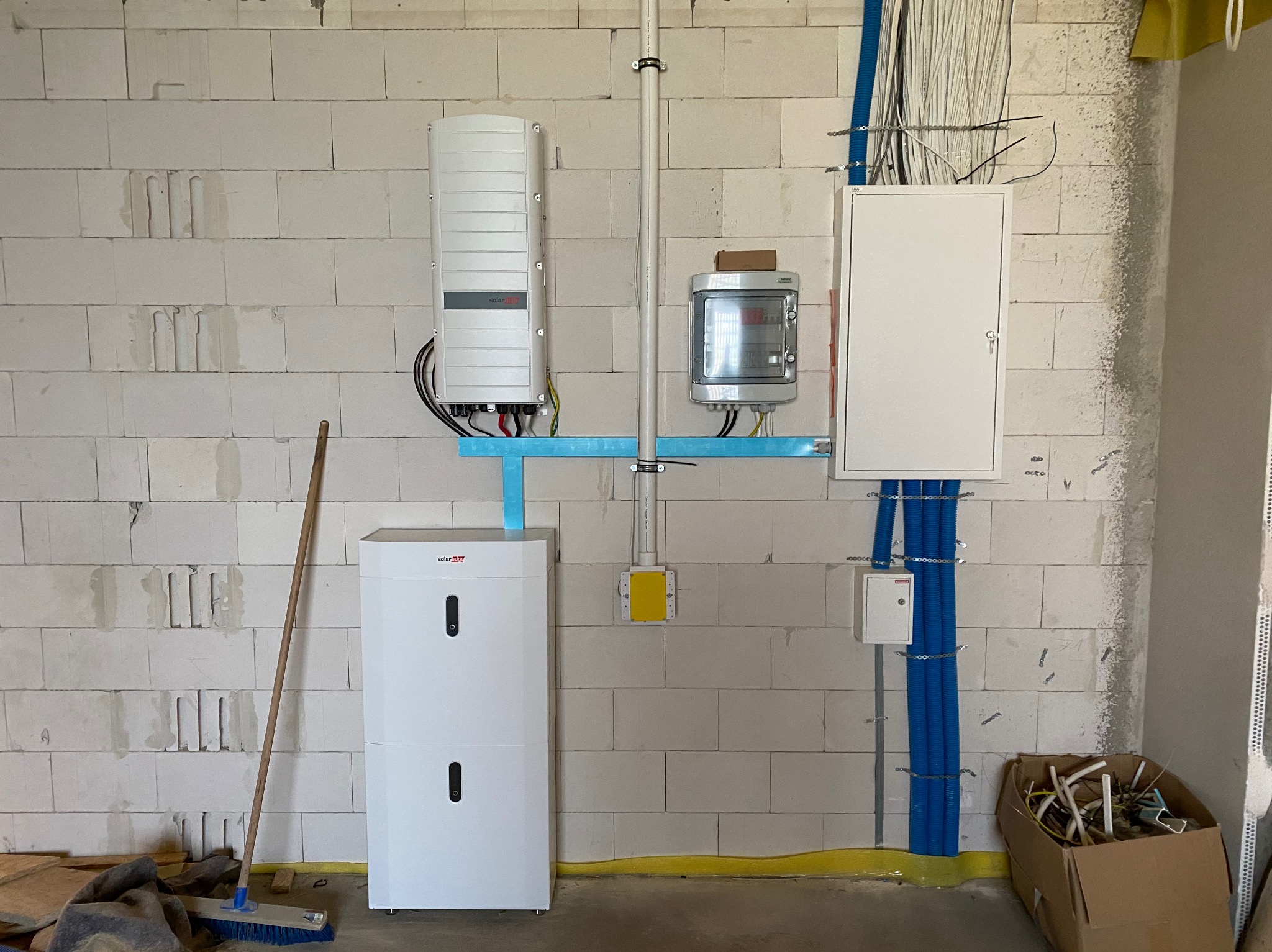 Biała skrzynka elektryczna, licznik i falownik na ścianie z betonu komórkowego. Niebieskie rurki osłonowe na kable. Widoczne przewody i elementy instalacji.