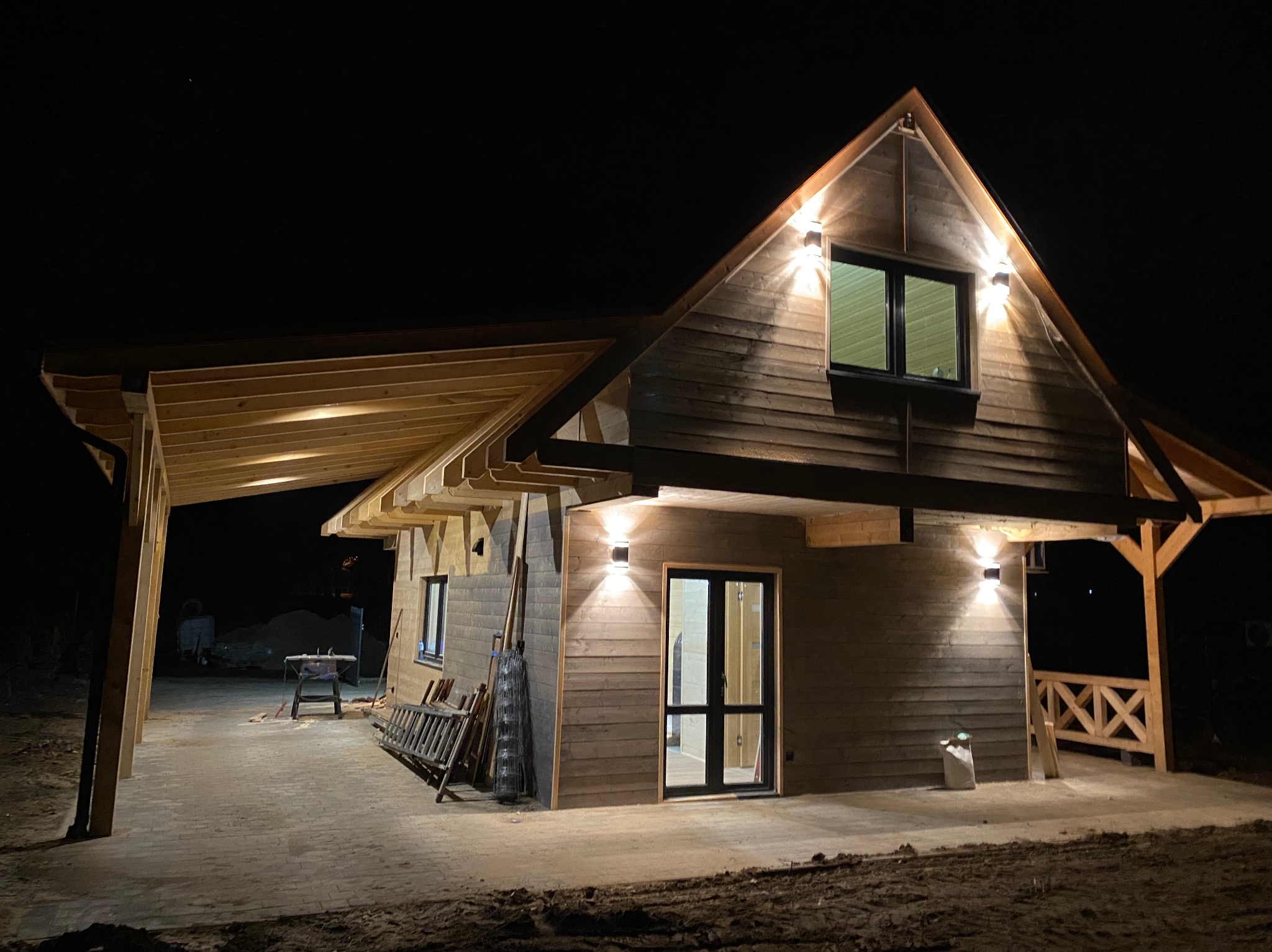 Nocny widok drewnianego domu z podświetleniem elewacji i zadaszonego tarasu. Widoczne lampy ścienne, okno i drzwi. Częściowo wykończony teren wokół budynku.