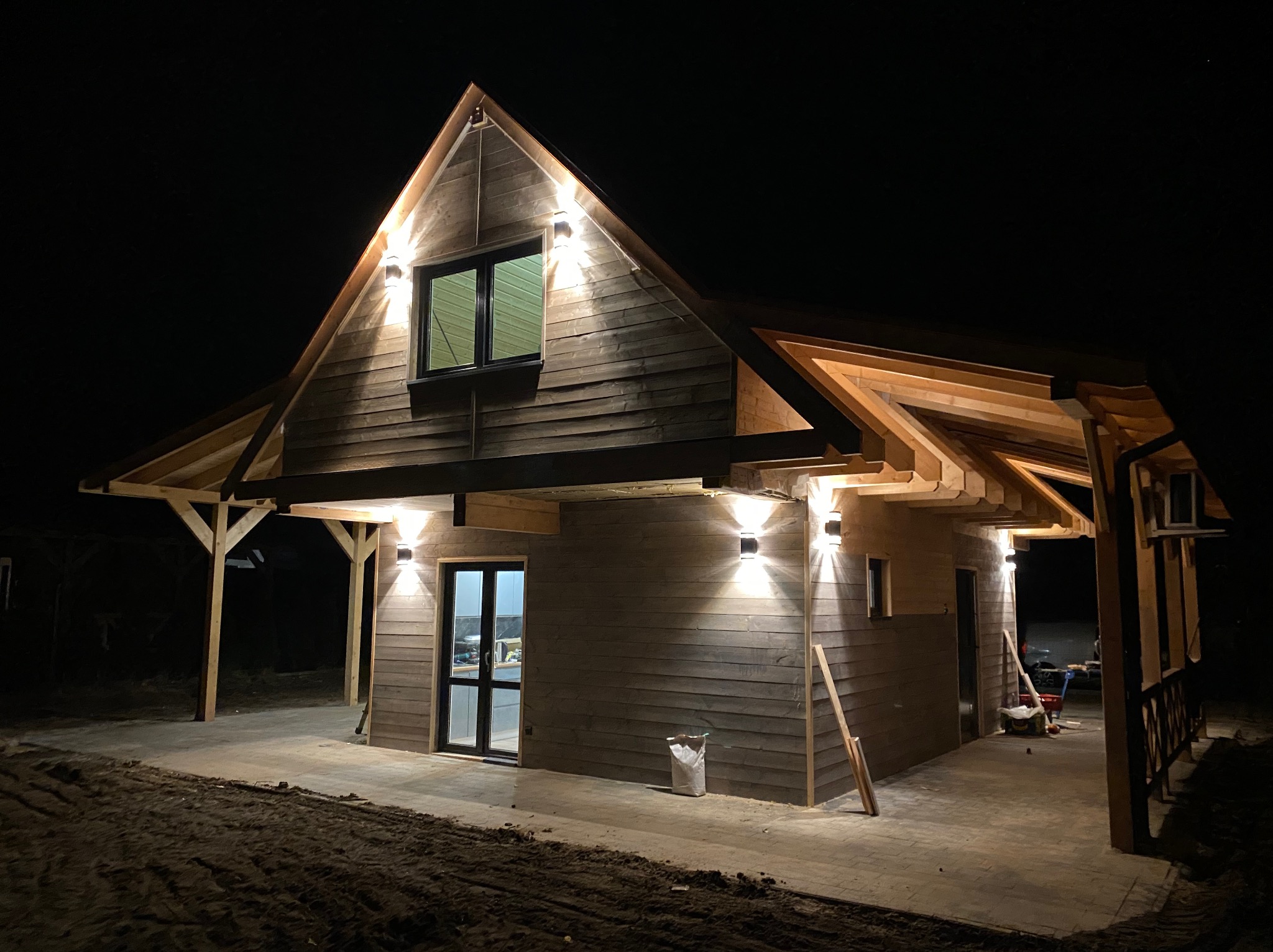 Nocny widok drewnianego domu z oświetleniem zewnętrznym. Lampy ścienne podkreślają architekturę budynku. Widoczne elementy konstrukcyjne dachu i tarasu.