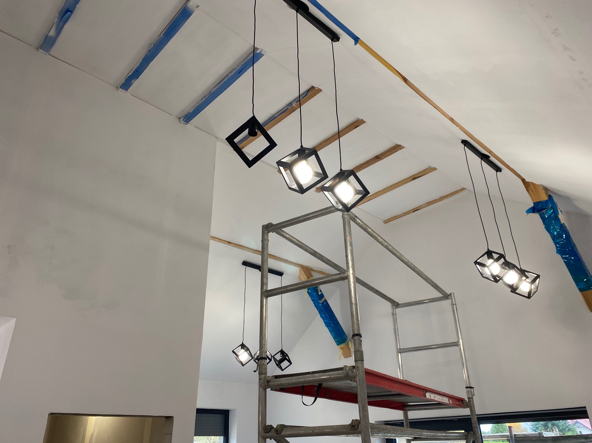 Montaż nowoczesnych lamp wiszących w surowym wnętrzu. Widoczne rusztowanie, zabezpieczenia malarskie i drewniane elementy konstrukcyjne. Oświetlenie w stylu industrialnym.