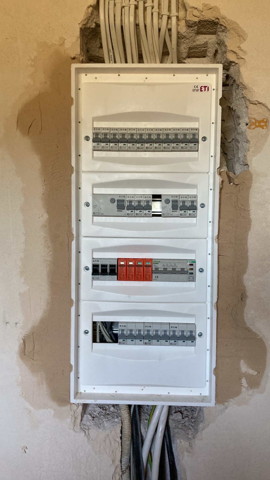 Rozdzielnica elektryczna z zabezpieczeniami w trakcie montażu, widoczne kable i fragment ściany z ubytkami tynku. Biała obudowa rozdzielnicy.