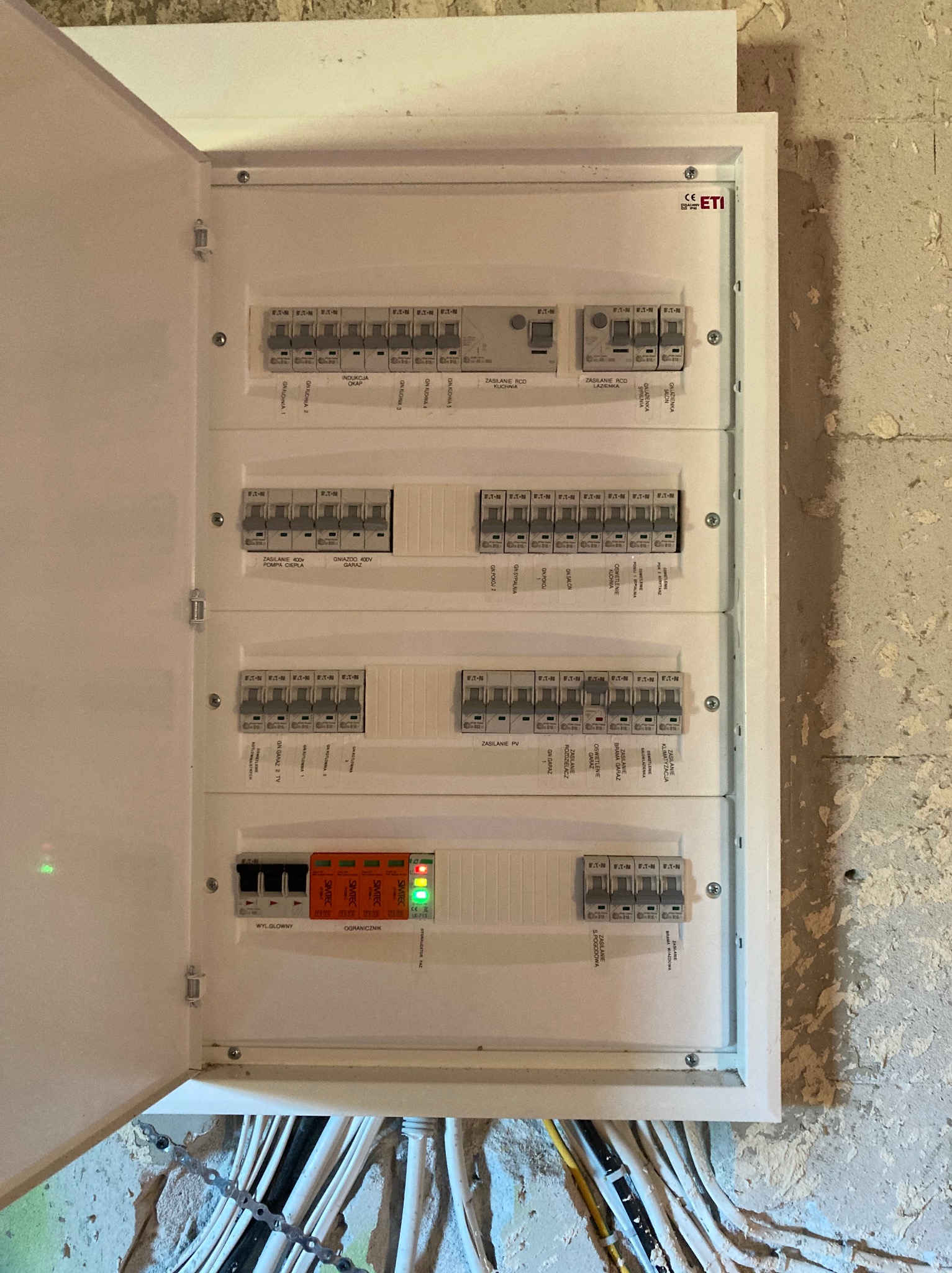 Otwarta skrzynka z bezpiecznikami ETI, widoczne zabezpieczenia przepięciowe SMITEC z diodami LED, opisane obwody i kable w ścianie.