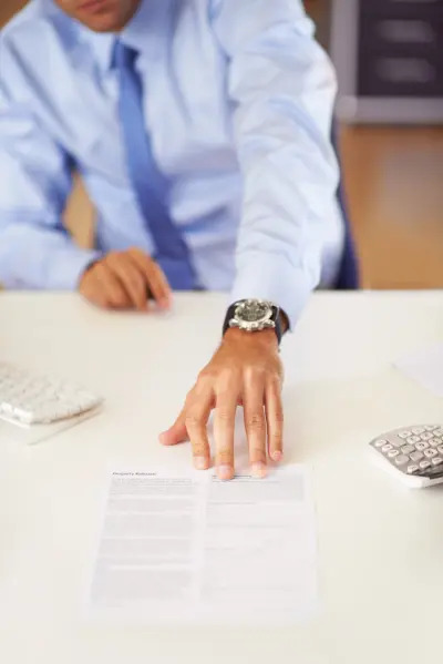 Mężczyzna w błękitnej koszuli i krawacie sięga ręką z zegarkiem po dokument leżący na białym biurku, obok klawiatury i kalkulatora.