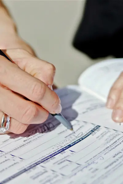 Ręka z pierścionkiem podpisuje dokument, prawdopodobnie wniosek lub umowę; widoczny fragment tekstu z polami do wypełnienia.