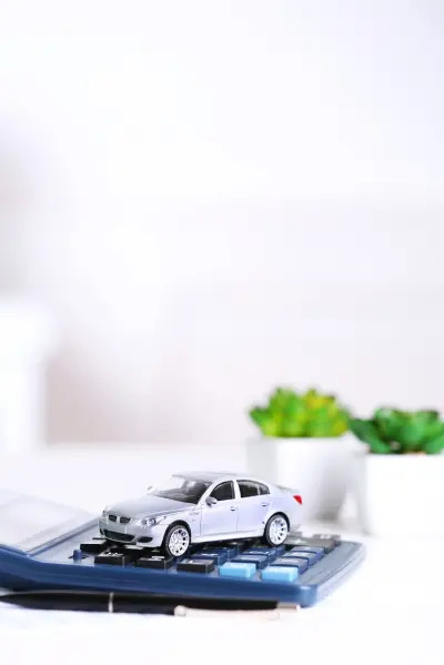 Miniaturowy srebrny samochód na kalkulatorze, obok doniczki z zieloną rośliną, na białym tle.