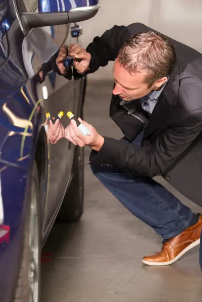 Mężczyzna w garniturze kuca obok niebieskiego samochodu, aplikując środek do usuwania rys z karoserii przy użyciu aplikatora z żółtą końcówką.