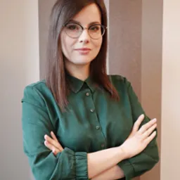 Portret kobiety w okularach i zielonej sukience, skrzyżowane ręce, neutralne tło w pionowe pasy. Uśmiech, profesjonalny wygląd.