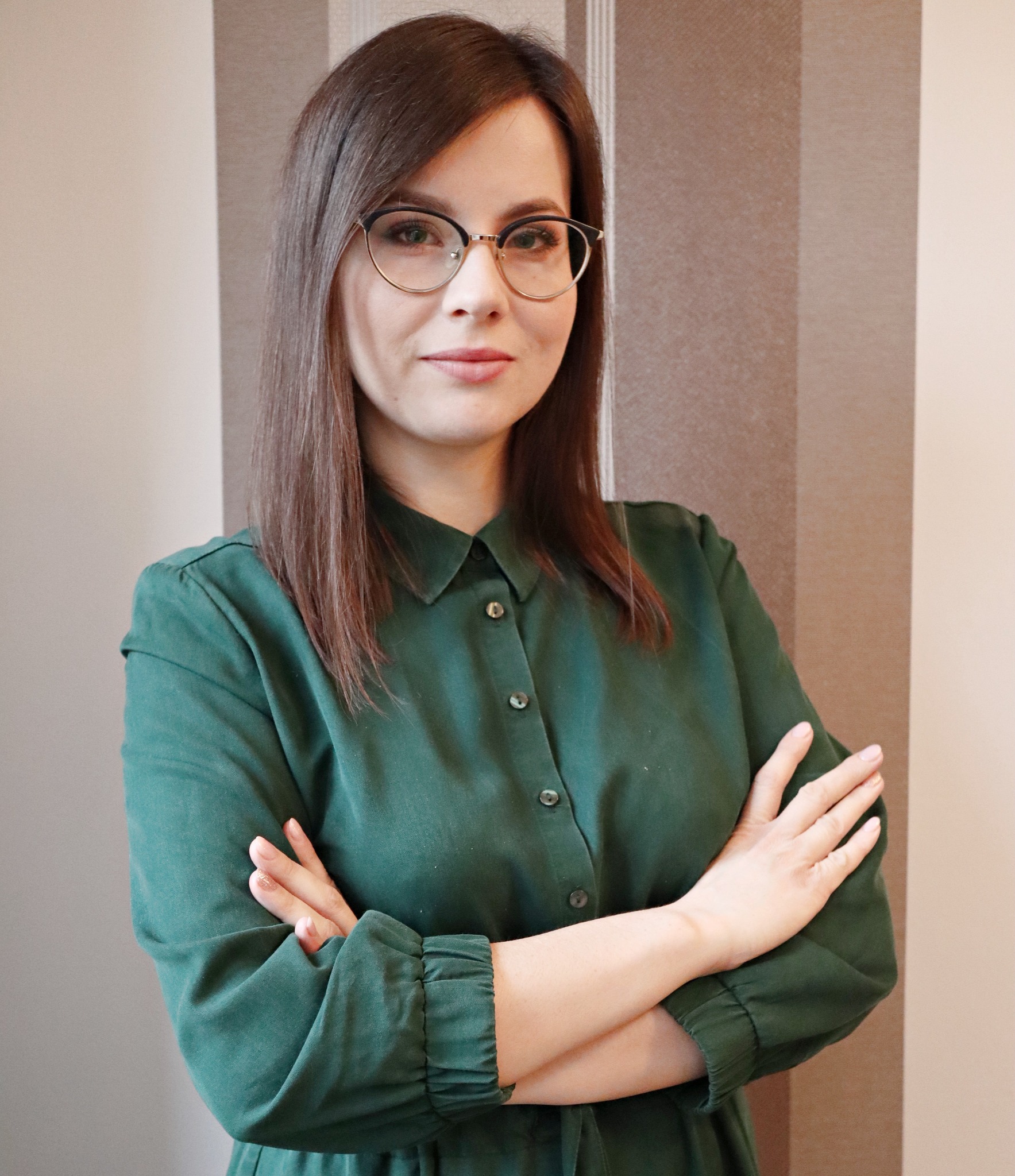 Portret kobiety w okularach i zielonej sukience, skrzyżowane ręce, neutralne tło w pionowe pasy. Uśmiech, profesjonalny wygląd.