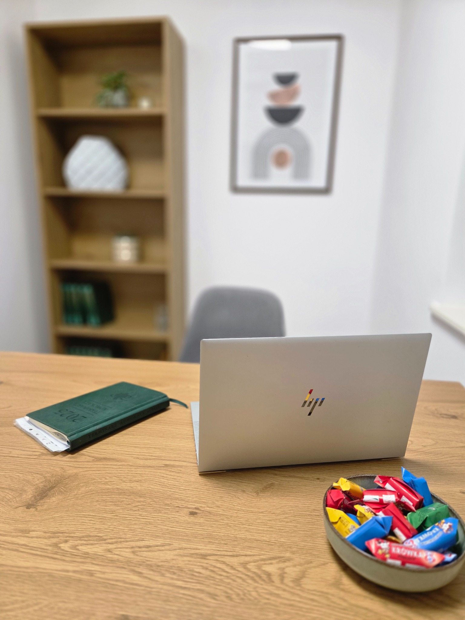 Biuro z laptopem, kalendarzem 2023 i miseczką cukierków na drewnianym biurku. W tle regał i grafika na ścianie. Jasne, neutralne kolory.