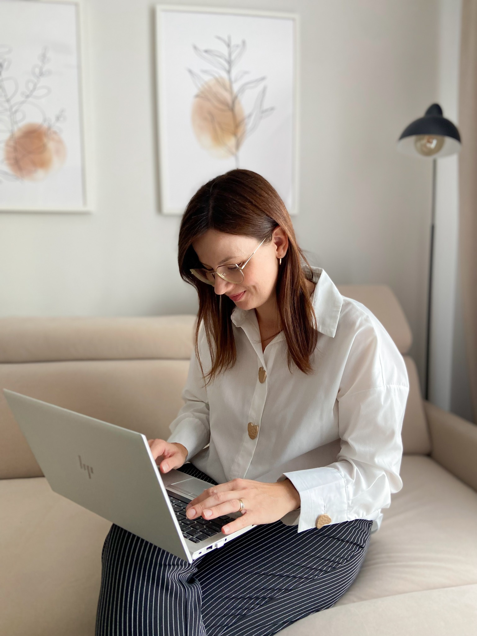 Kobieta w okularach, w białej koszuli z ozdobnymi guzikami, siedzi na beżowej sofie i pracuje na laptopie. W tle obrazy i lampa.
