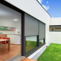 Nowoczesny dom z dużymi oknami osłoniętymi szarymi roletami zewnętrznymi, widok z wnętrza na zadbany trawnik i błękitne niebo.