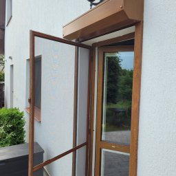 Plisy M&M - Otwarta moskitiera ramkowa w kolorze brązowym, dopasowanym do ramy okiennej i obudowy rolety zewnętrznej, zamontowana na elewacji domu z widoczną lampą zewnętrzną.