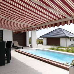 Plisy M&M - Zadaszony taras z basenem, chroniony rozsuwaną markizą w biało-bordowe pasy, z widokiem na zadbany ogród i budynek garażowy.