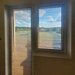 Jasne, drewniane żaluzje poziome w oknie, częściowo zasłonięte, z widokiem na zewnątrz w słoneczny dzień.