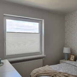 Plisy M&M - Jasna plisa okienna w sypialni z kwiatową tapetą. Widok na niebo przez okno. Grzejnik pod oknem. Dekoracje na stole. Stonowana kolorystyka.