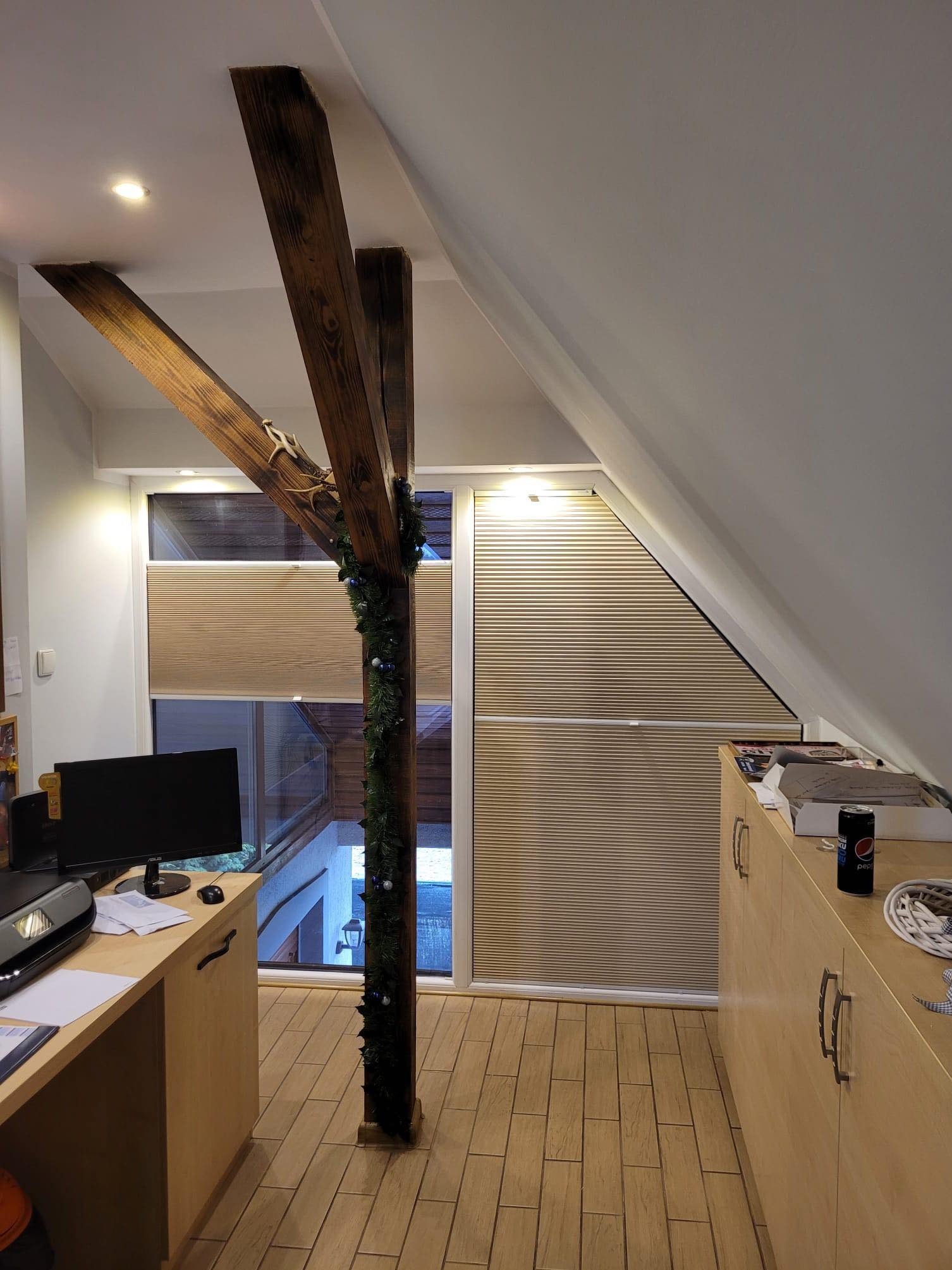 Beżowe żaluzje plisowane, dopasowane do okna o nietypowym, trójkątnym kształcie na poddaszu, widoczne drewniane elementy konstrukcyjne pomieszczenia, w tle biurko z komputerem.