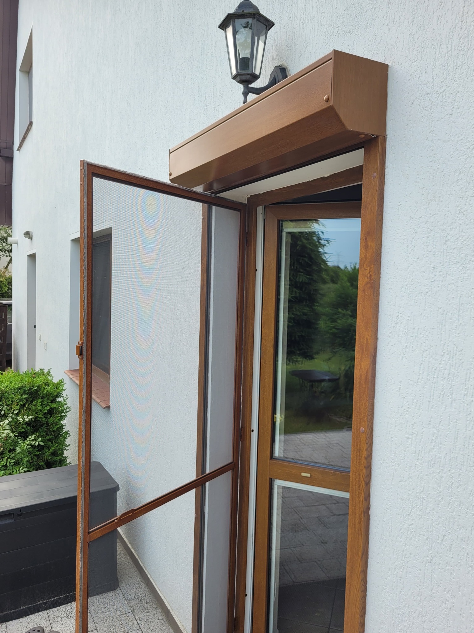 Otwarta moskitiera ramkowa w kolorze brązowym, dopasowanym do ramy okiennej i obudowy rolety zewnętrznej, zamontowana na elewacji domu z widoczną lampą zewnętrzną.