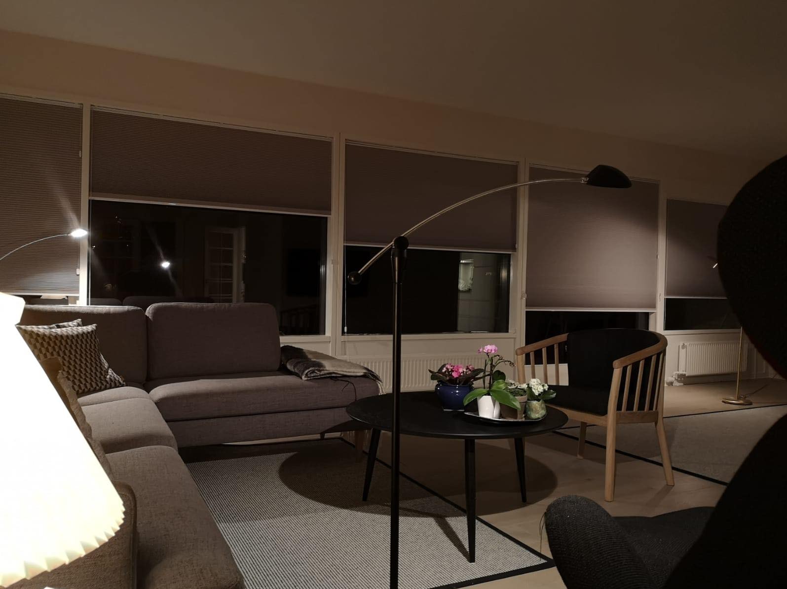 Zasłonięte okna w salonie z szarymi roletami plisowanymi, widoczne przytulne wnętrze z sofą, stolikiem i lampą łukową.