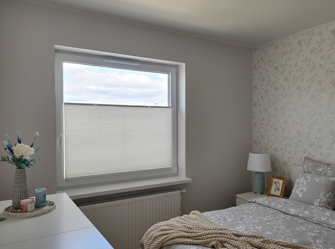 Jasna plisa okienna w sypialni z kwiatową tapetą. Widok na niebo przez okno. Grzejnik pod oknem. Dekoracje na stole. Stonowana kolorystyka.