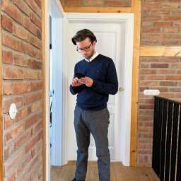 PAWEŁ TYL NIERUCHOMOŚCI - Mężczyzna w okularach, stojący w korytarzu z ceglanymi ścianami i drewnianą podłogą, przegląda coś na telefonie.