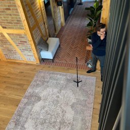 PAWEŁ TYL NIERUCHOMOŚCI - Wnętrze domu z widokiem z góry: parkiet, dywan w stylu vintage, ceglana ściana z drewnianą konstrukcją, mężczyzna w okularach oparty o balustradę.