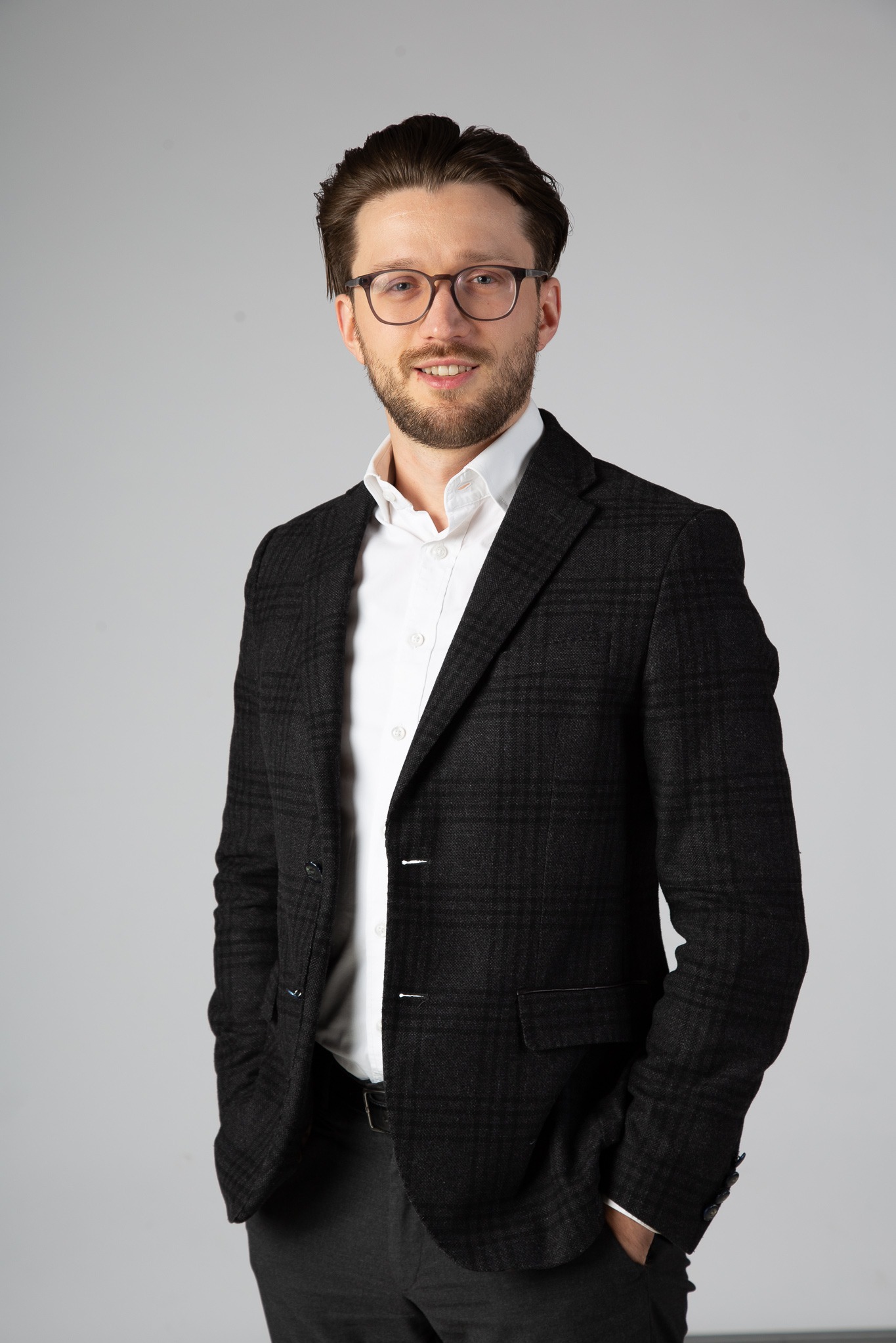 Portret uśmiechniętego mężczyzny w okularach, białej koszuli i ciemnej marynarce w kratę na jasnoszarym tle.