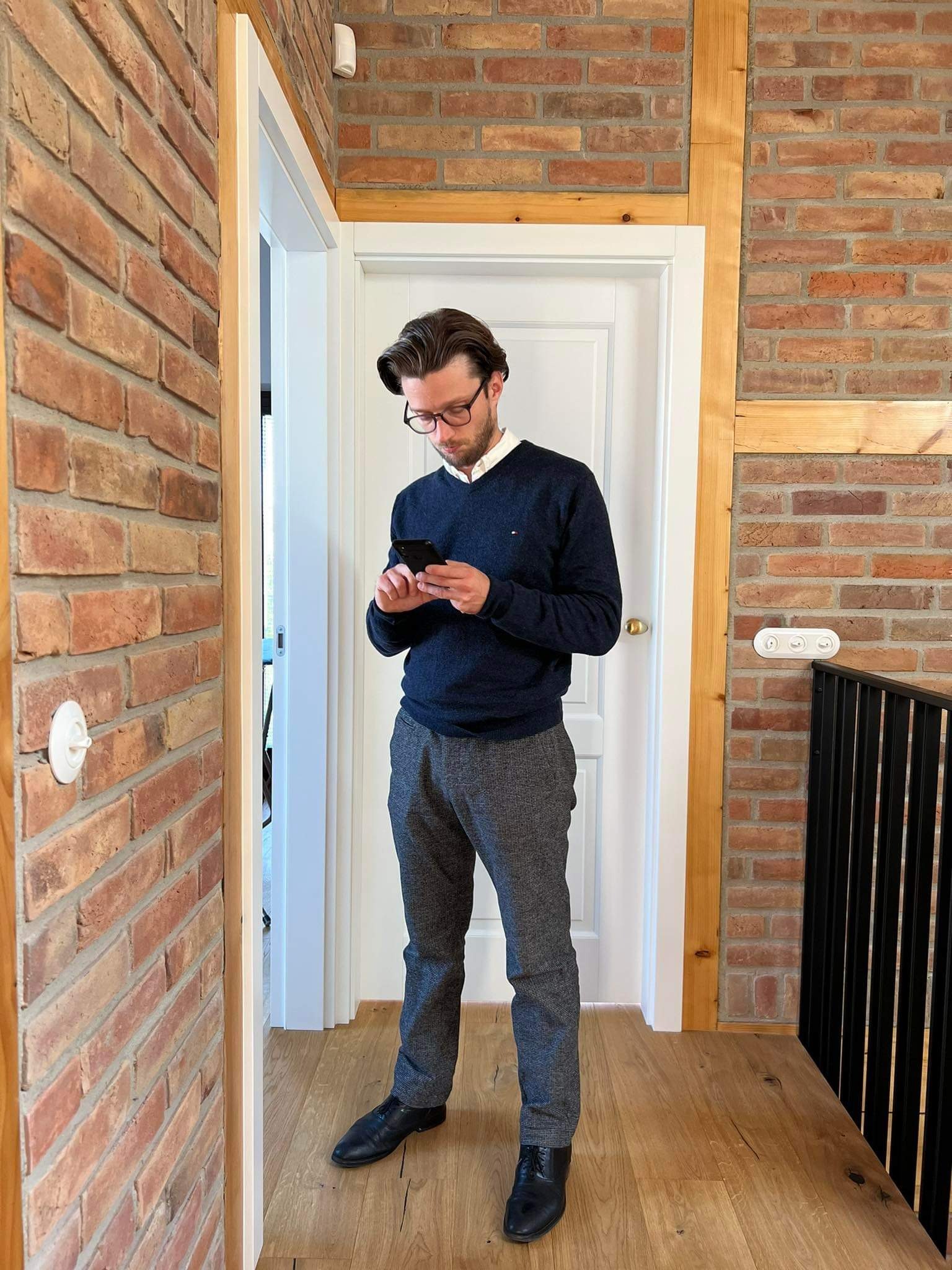 Mężczyzna w okularach, stojący w korytarzu z ceglanymi ścianami i drewnianą podłogą, przegląda coś na telefonie.
